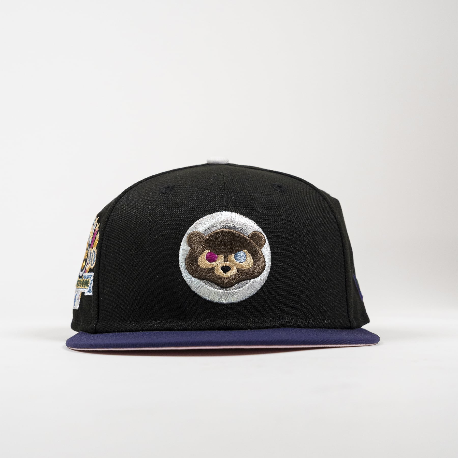 A black flat-brim cap with a dark purple brim, featuring a circular embroidered patch of a raccoon wearing a space helmet on the front. The side of the cap has a colorful patch.