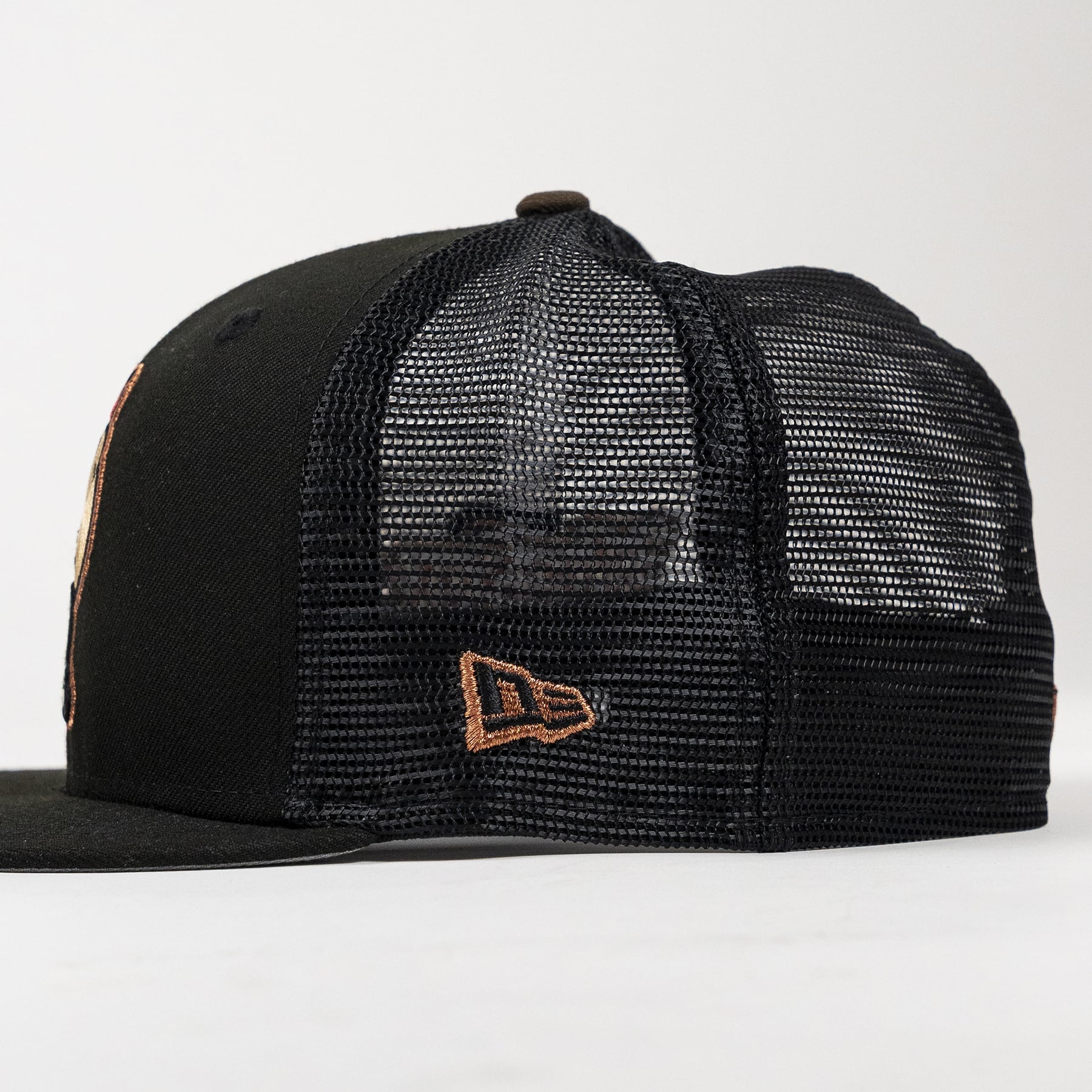 A black trucker-style baseball cap with a black mesh back and a small brown embroidered New Era logo on the side, shown against a plain light background.