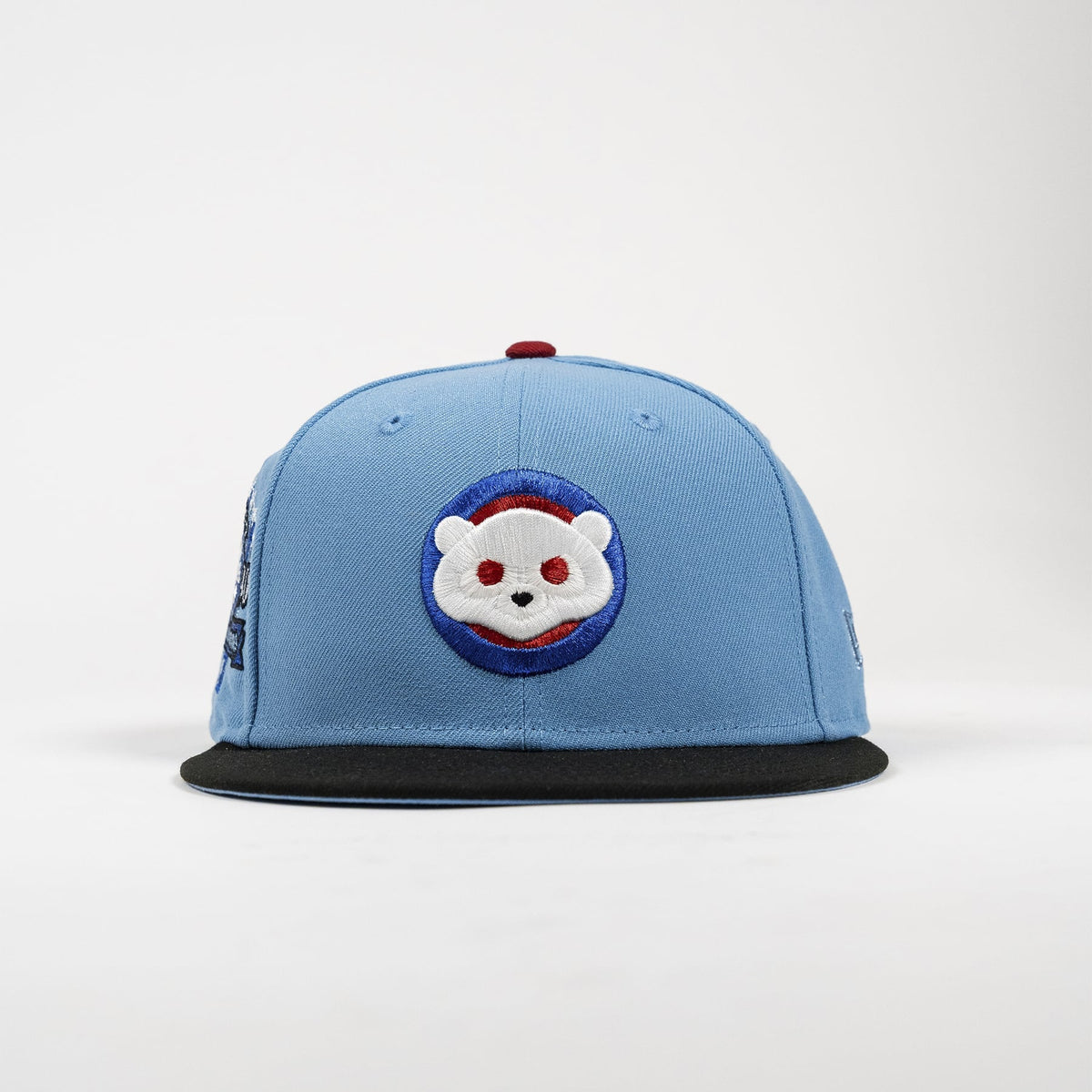 A light blue flat-brim cap featuring a circular embroidered patch of a white cartoon bear face with red eyes on a blue and red background, with a black visor and a small red button on top.