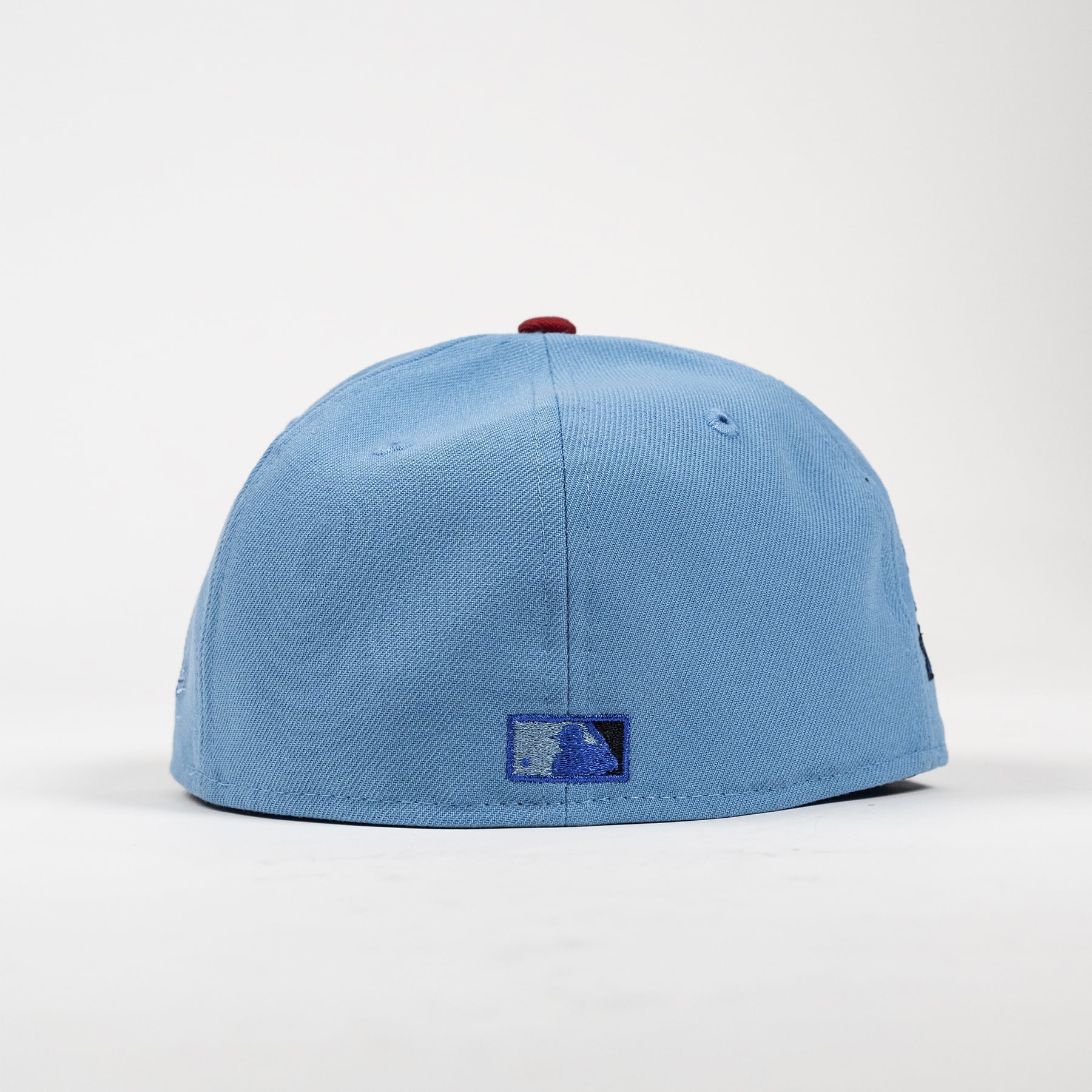 A light blue baseball cap with a small red button on top and a blue embroidered patch on the front, photographed against a clean white background.