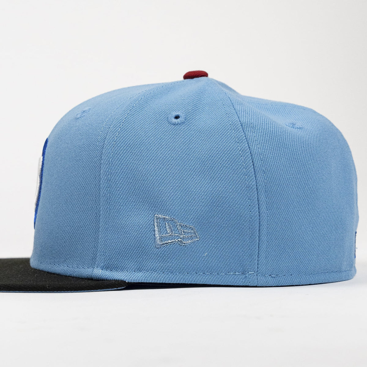 A light blue structured baseball cap with a black flat brim, featuring a silver embroidered New Era logo on the front and a small red button on top, photographed on a white background.