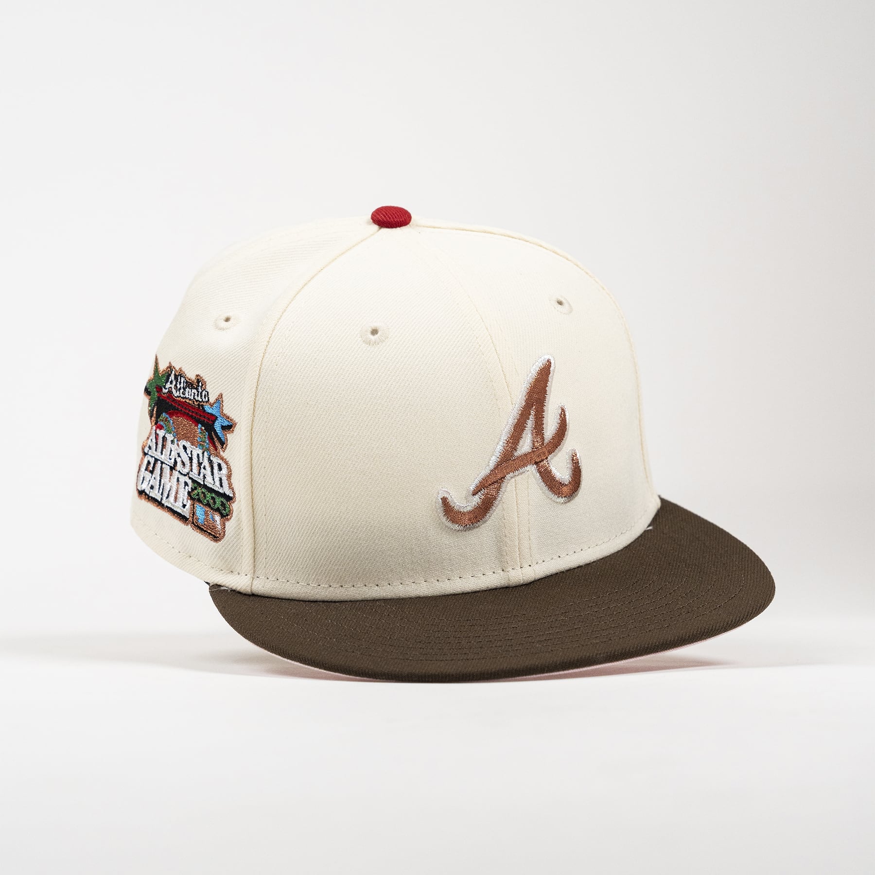 An off-white baseball cap with a brown flat brim, featuring a brown embroidered Atlanta 'A' logo on the front and a '2024 All-Star Game' patch on the side. The cap has a small red button on top and is photographed on a clean white background.