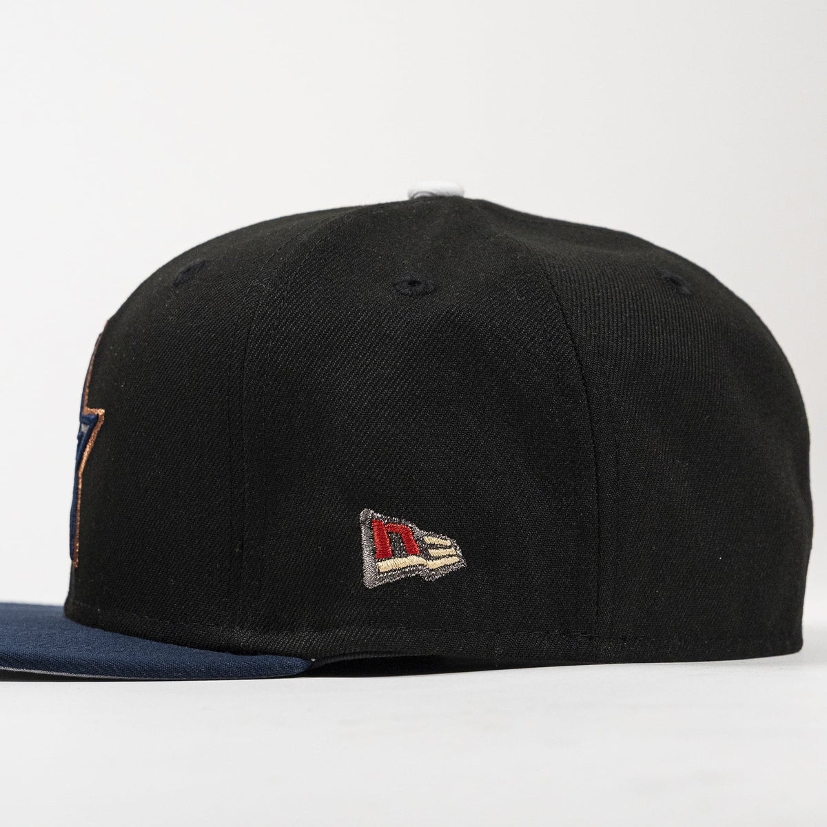 A black structured baseball cap with a navy blue flat brim, featuring a small embroidered New Era flag logo on the side and a partial embroidered patch on the opposite side.