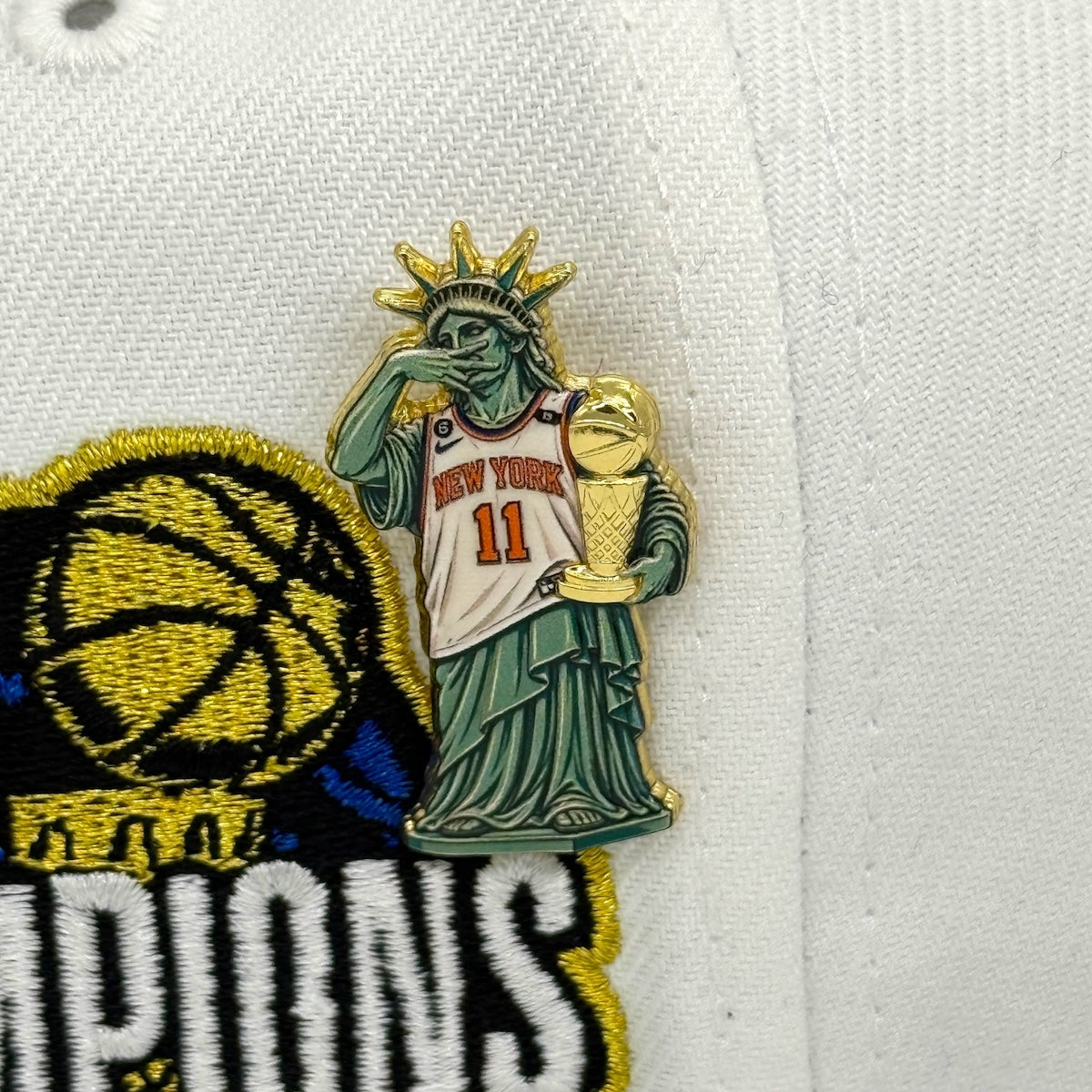 A close-up of a white baseball cap featuring a green and gold enamel pin of a Statue of Liberty dressed in a New York Knicks jersey with the number 11, next to a black, yellow, and white embroidered patch.