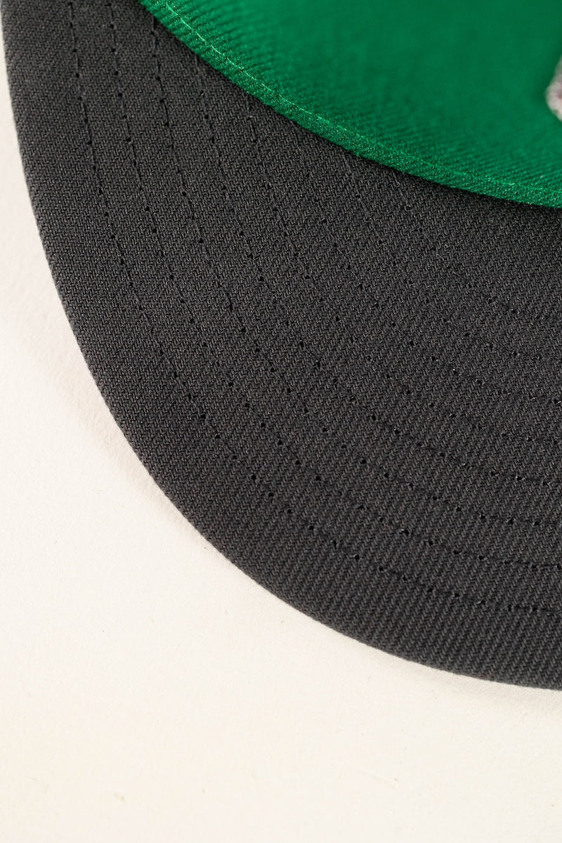 Close-up detail shot of a snapback cap showing the black curved brim with stitched edge and a green fabric crown visible at the top.
